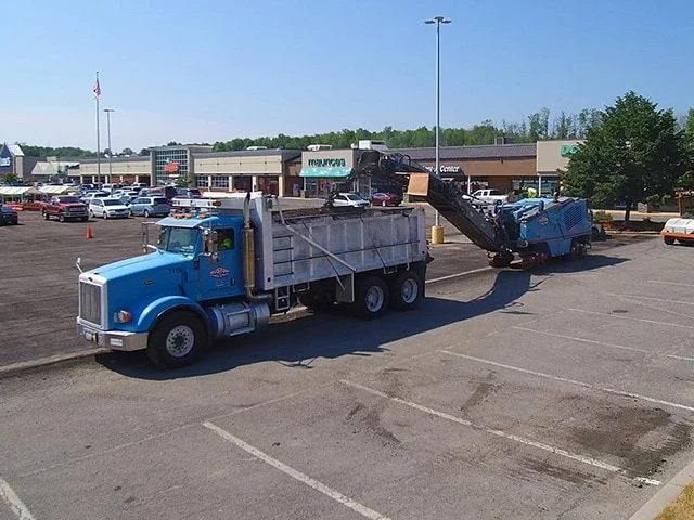 Asphalt milling machine removing old pavement in a commercial parking lot while loading milled asphalt into a dump truck for recycling during asphalt resurfacing project